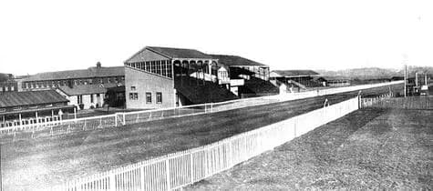 A grayscale photo depicts a long, straight race track with a white fence running along its edge, and grandstands and other buildings are visible in the background under a clear sky.