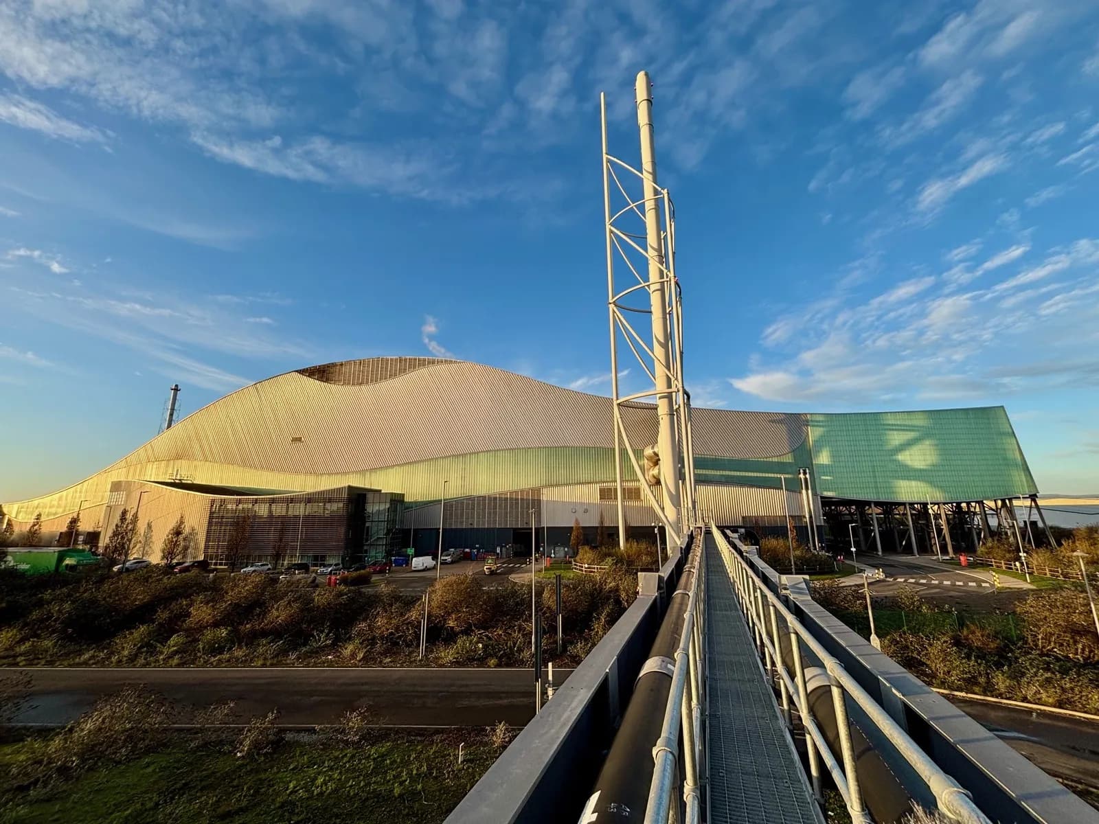 A wide shot shows an expansive, modern building with a curved, light-colored roof and green-paneled sections under a blue sky with scattered clouds, with a tall, white industrial structure and a walkway in the foreground.